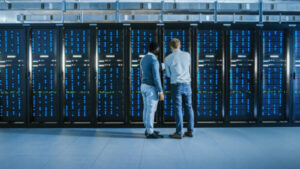 Bearded IT Technician in Glasses with a Laptop Computer and Black Male Engineer Colleague are Talking in Data Center while Working Next to Server Racks. Running Diagnostics or Doing Maintenance Work.